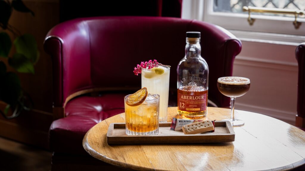 Three shortbread whisky cocktails on a wooden table at The Craigellachie Hotel, with an Aberlour whisky bottle and Walker's Shortbread Finger, set against the warm leather chairs of The Quaich Bar.